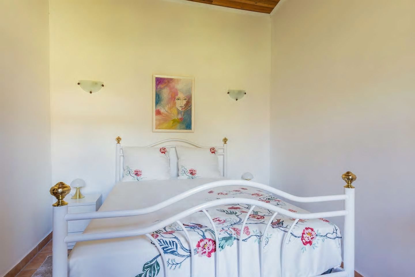 A bedroom with a white bed, floral bedspread, and a painting on the wall.