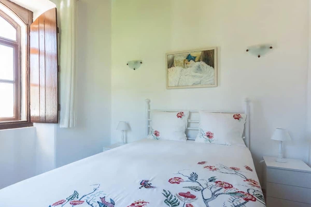 A bedroom with a bed, two bedside tables, a window with curtains, and a painting on the wall.