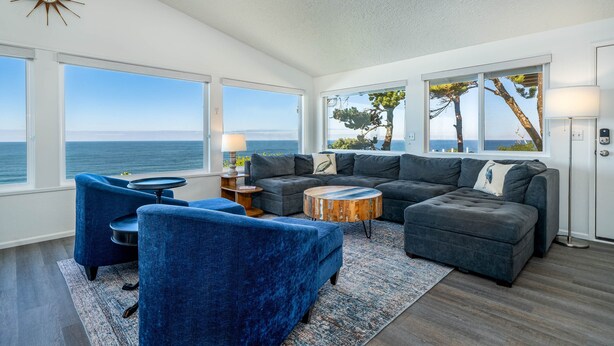 A modern living room with a large window offering a view of the sea, a cozy seating area with blue sofas, and a wooden coffee table. - Take in panoramic views from the living room or deck at this oceanfront home (Lincoln City)