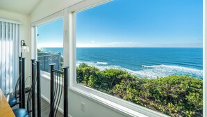 A room with a large window overlooking the ocean, a wooden table, and black metal railings. - Take in panoramic views from the living room or deck at this oceanfront home (Lincoln City)
