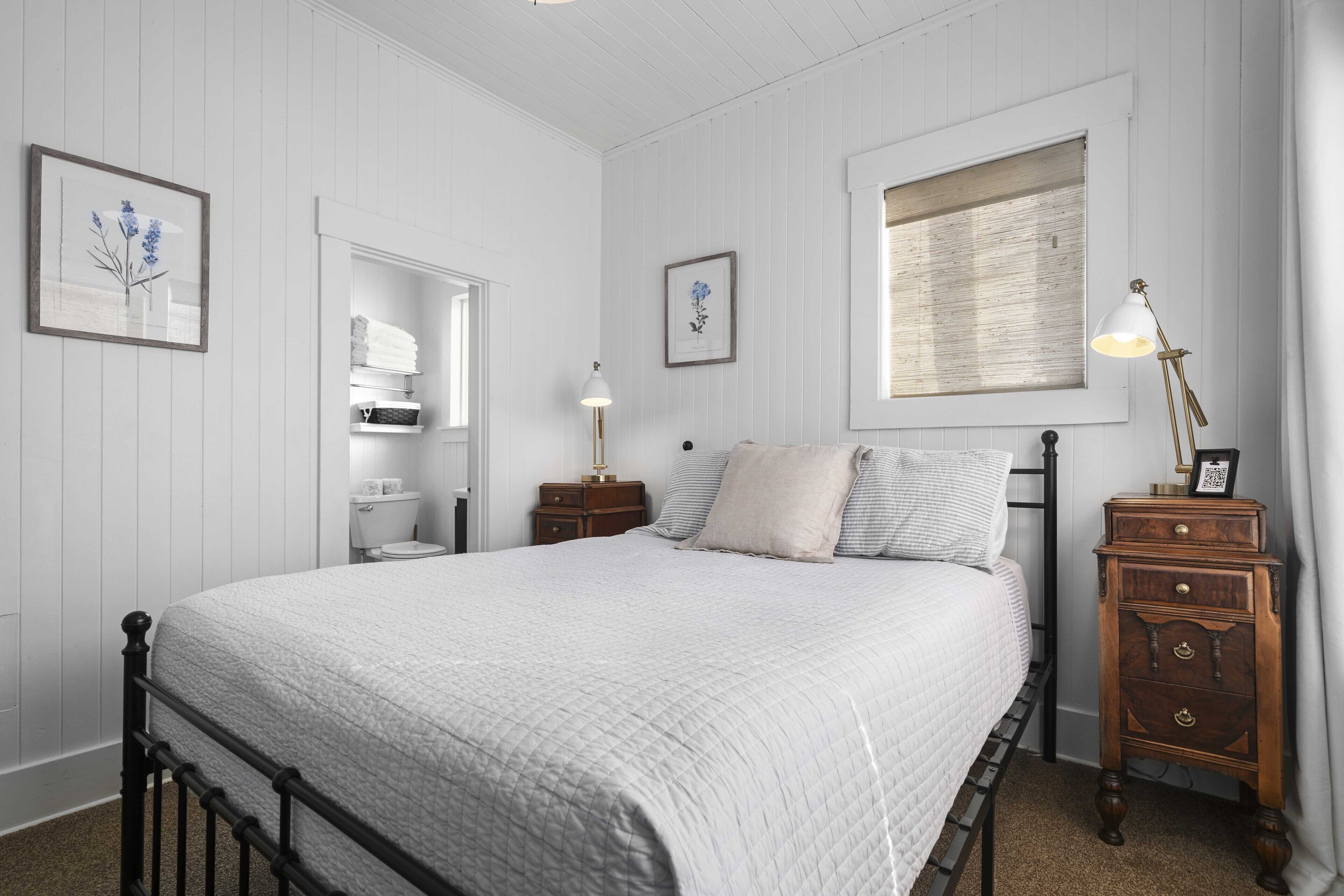 A bedroom with a bed, nightstands, a dresser, a window with blinds, and framed pictures on the walls.