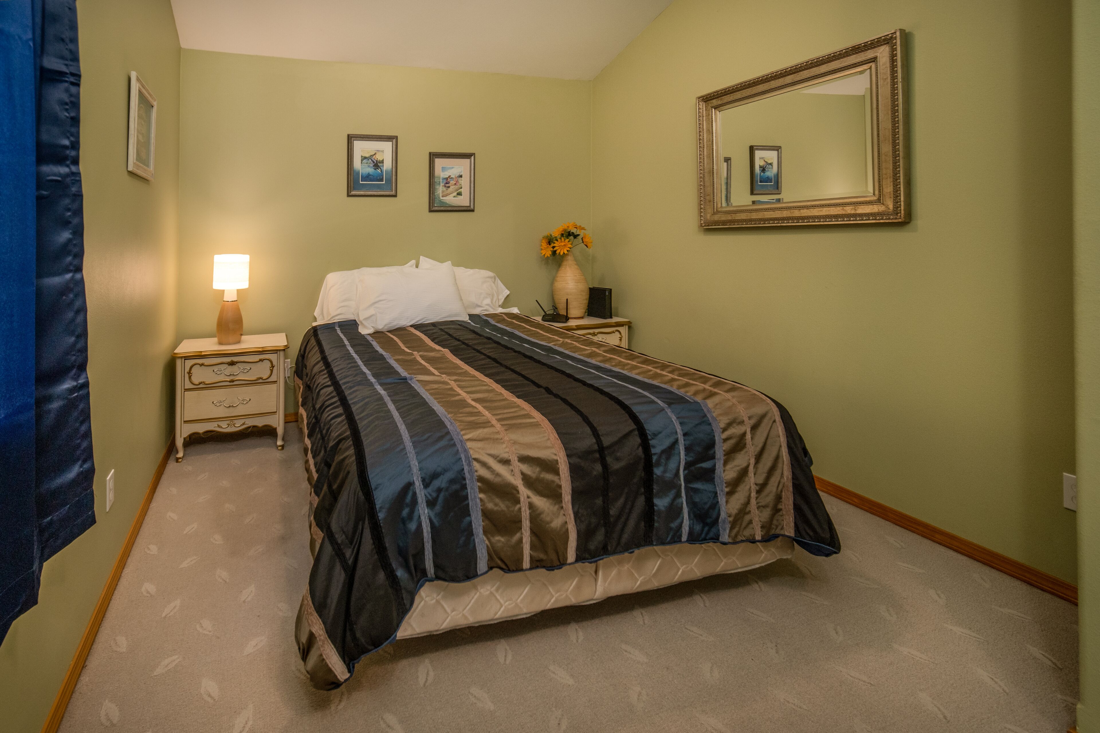A bedroom with a bed, a nightstand, a lamp, a vase with flowers, and framed pictures on the wall.