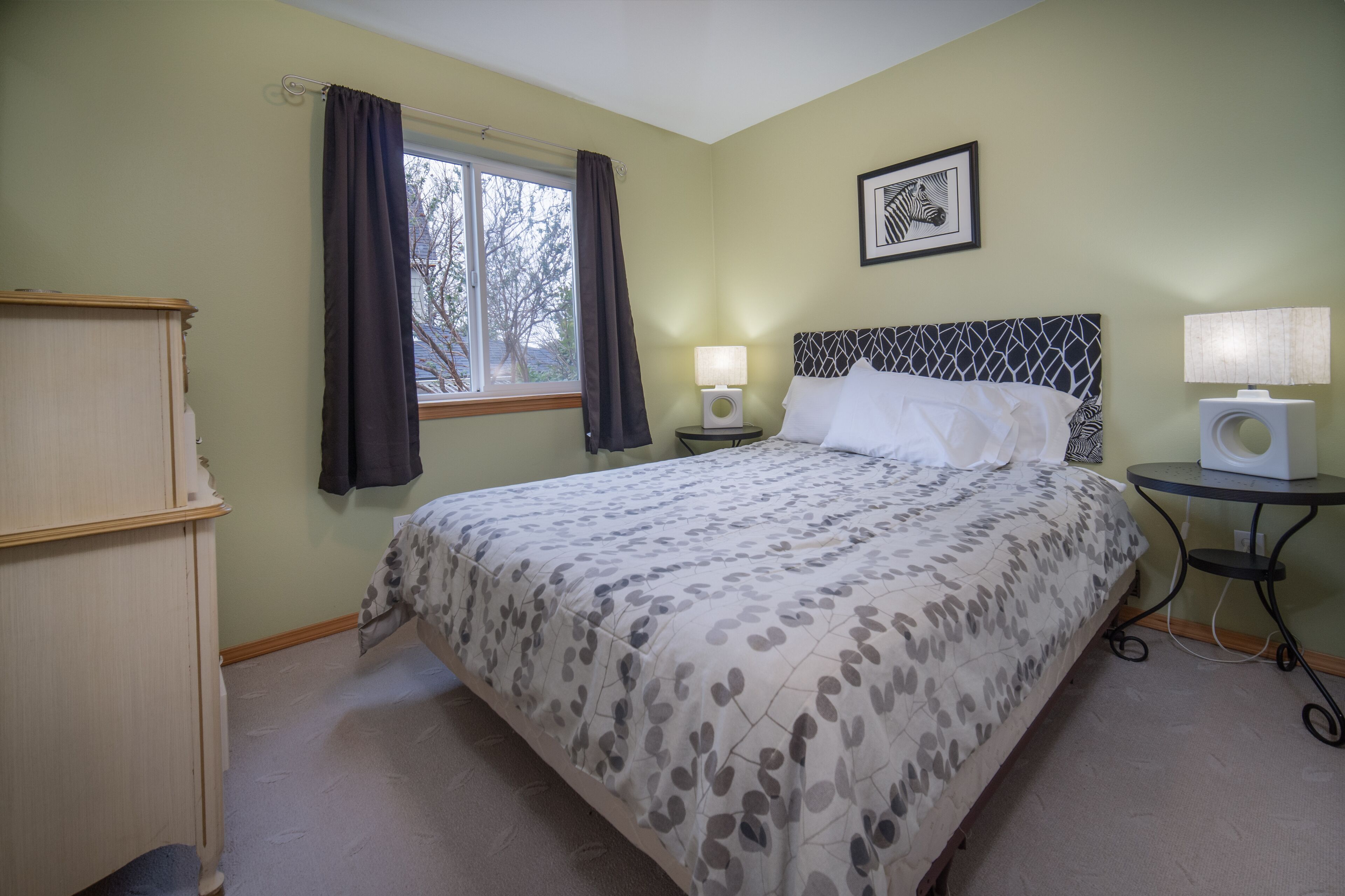 A bedroom with a bed, two bedside lamps, a window with curtains, a framed picture on the wall, and a small table with a lamp.