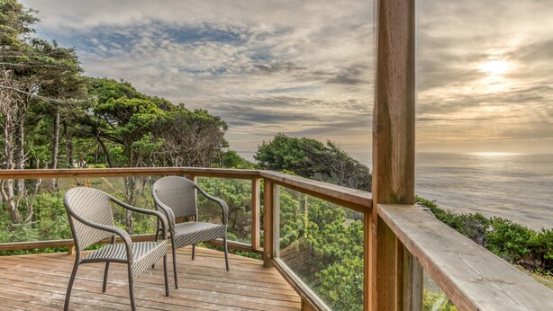 Eine Holzterrasse mit zwei Korbstühlen mit Blick auf eine Küstenlandschaft mit Bäumen und dem Meer.