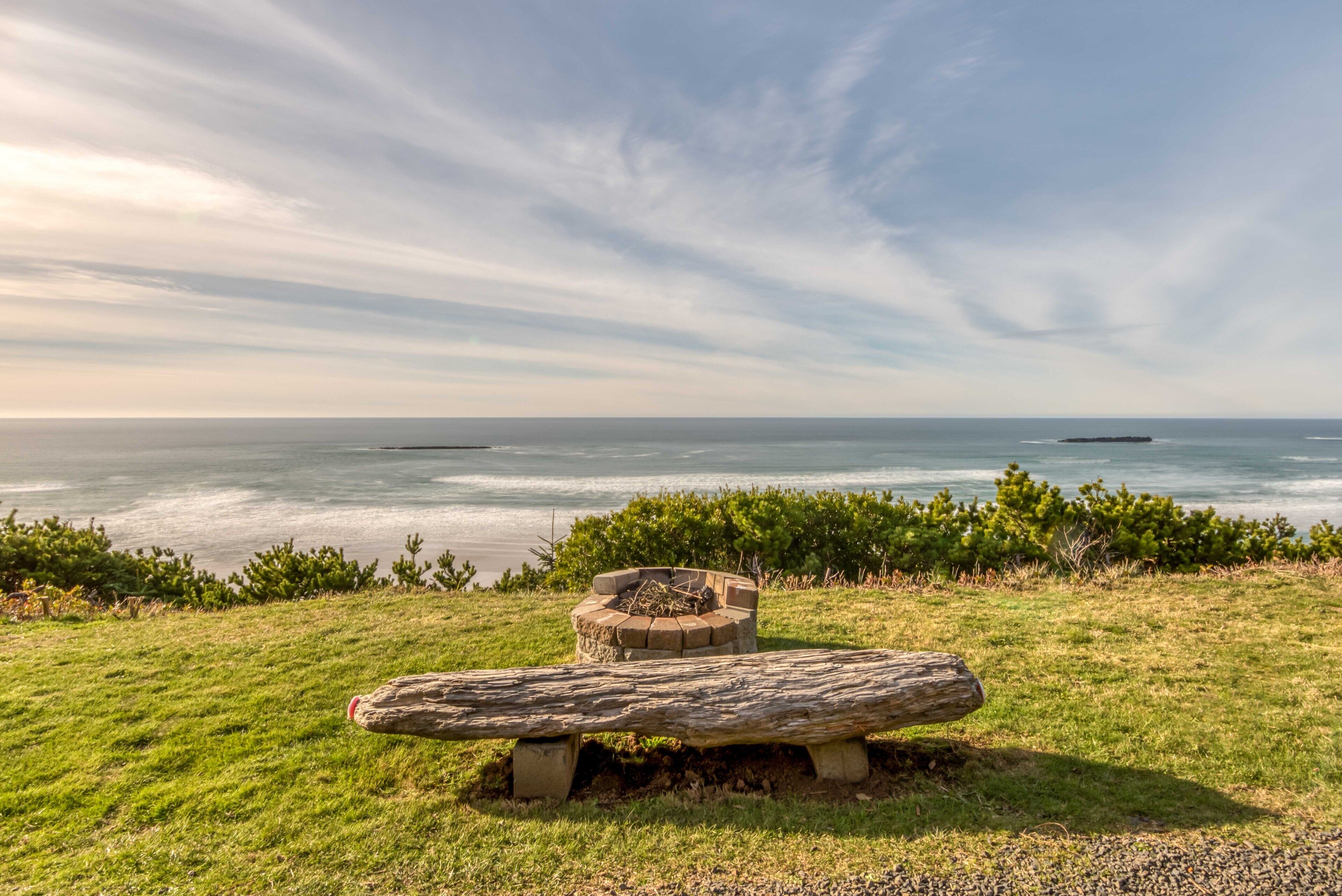 A bench and a fire pit on a grassy hill overlooking a calm sea with distant islands.