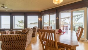 Un comedor con una mesa y sillas de madera, ventanas amplias con vistas a un puente, y sofás estampados.