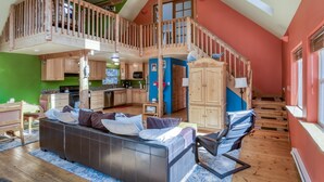 A spacious loft with a high ceiling, a wooden staircase, a kitchen with wooden cabinets, a dining area with a sofa, and a large window providing natural light. - Pet Friendly Home on a Secluded Wooded Lot only 4 Blocks to the Beach (Yachats)