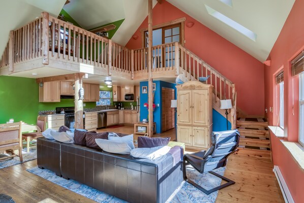 Un loft espacioso con techo alto, una escalera de madera, cocina con armarios de madera, zona de comedor con sofá y un ventanal que proporciona luz natural.