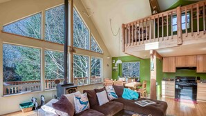 A spacious living area with a large brown sofa, wooden dining table, and a kitchen with green cabinets. - Pet Friendly Home on a Secluded Wooded Lot only 4 Blocks to the Beach (Yachats)