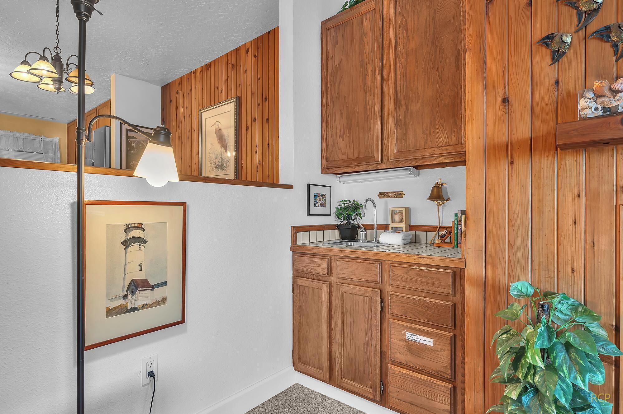 A kitchen with wooden cabinets, a sink, and a pendant light. There are framed pictures on the wall and a plant on the counter.