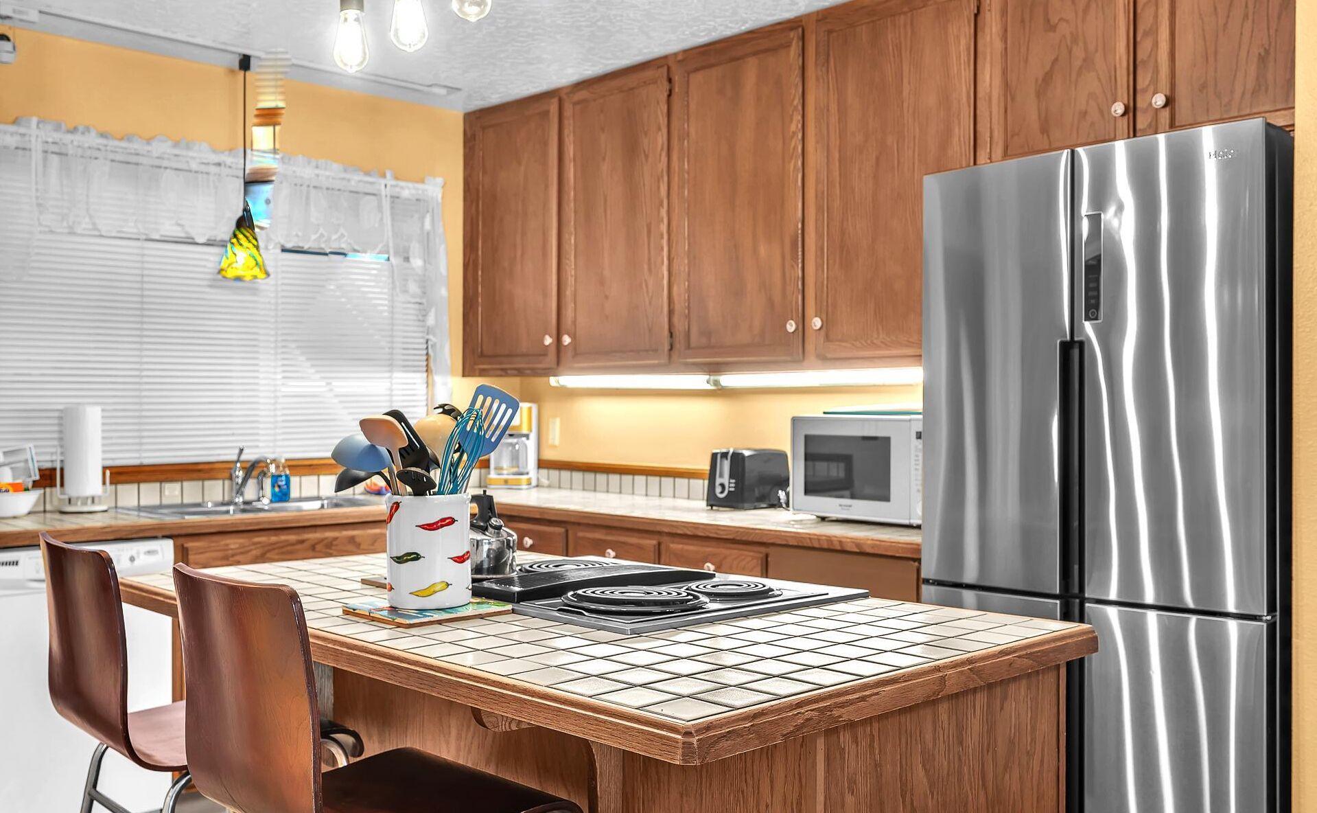 A modern kitchen with wooden cabinets, a stainless steel refrigerator, a microwave, and a tiled island with chairs.