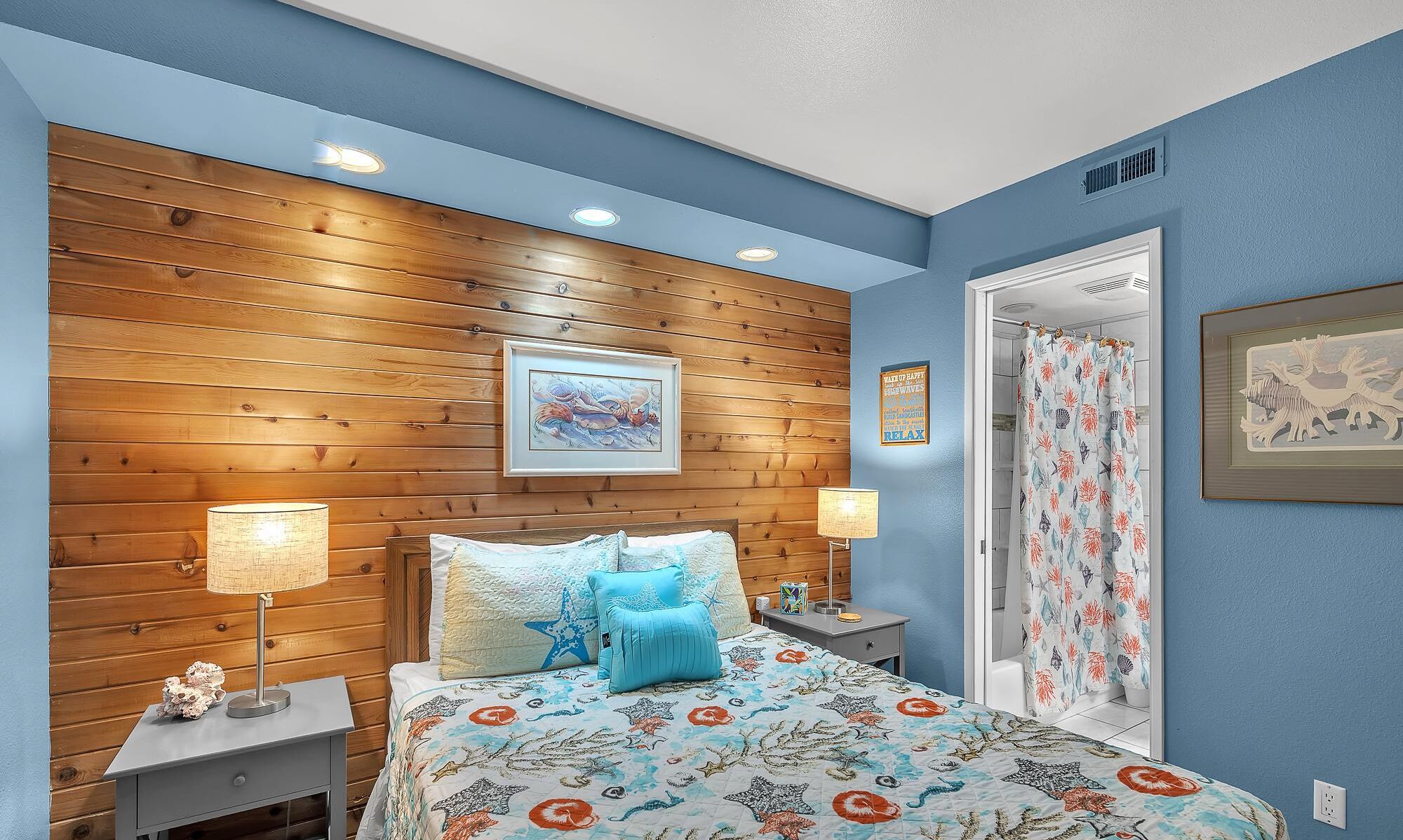 A bedroom with a wooden headboard, a bed with a floral bedspread, two bedside lamps, and a framed picture above the bed.