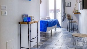 A compact hotel room with a bed, a small table, a chair, and a blue-striped pet bed. - B&B Fico d'India (Polignano a Mare)