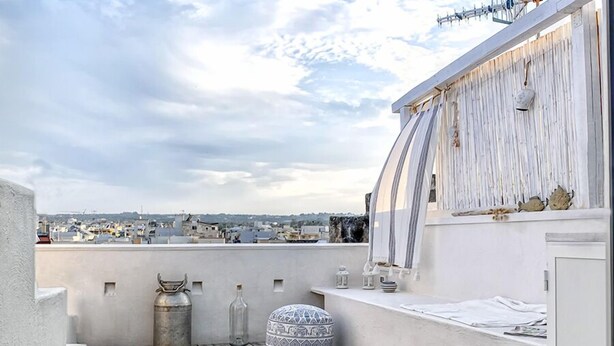 A rooftop terrace with a wooden floor, a metal gas cylinder, a patterned cushion, and a white wall with a window. - B&B Fico d'India (Polignano a Mare)