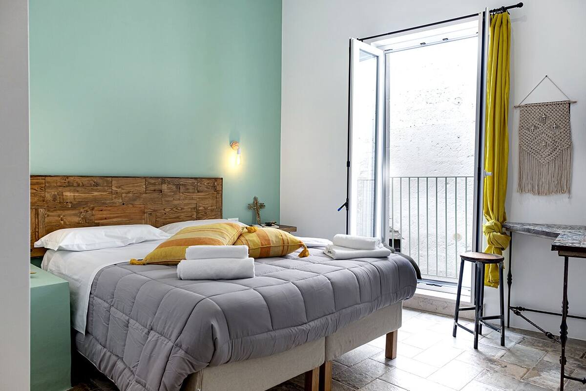 A bedroom with a bed, a nightstand, a desk, and a window with yellow curtains.