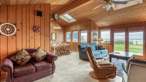 Een ruime woonkamer met een houten plafond, een blauwe bank, een bruine fauteuil en een eethoek met een houten tafel en stoelen.