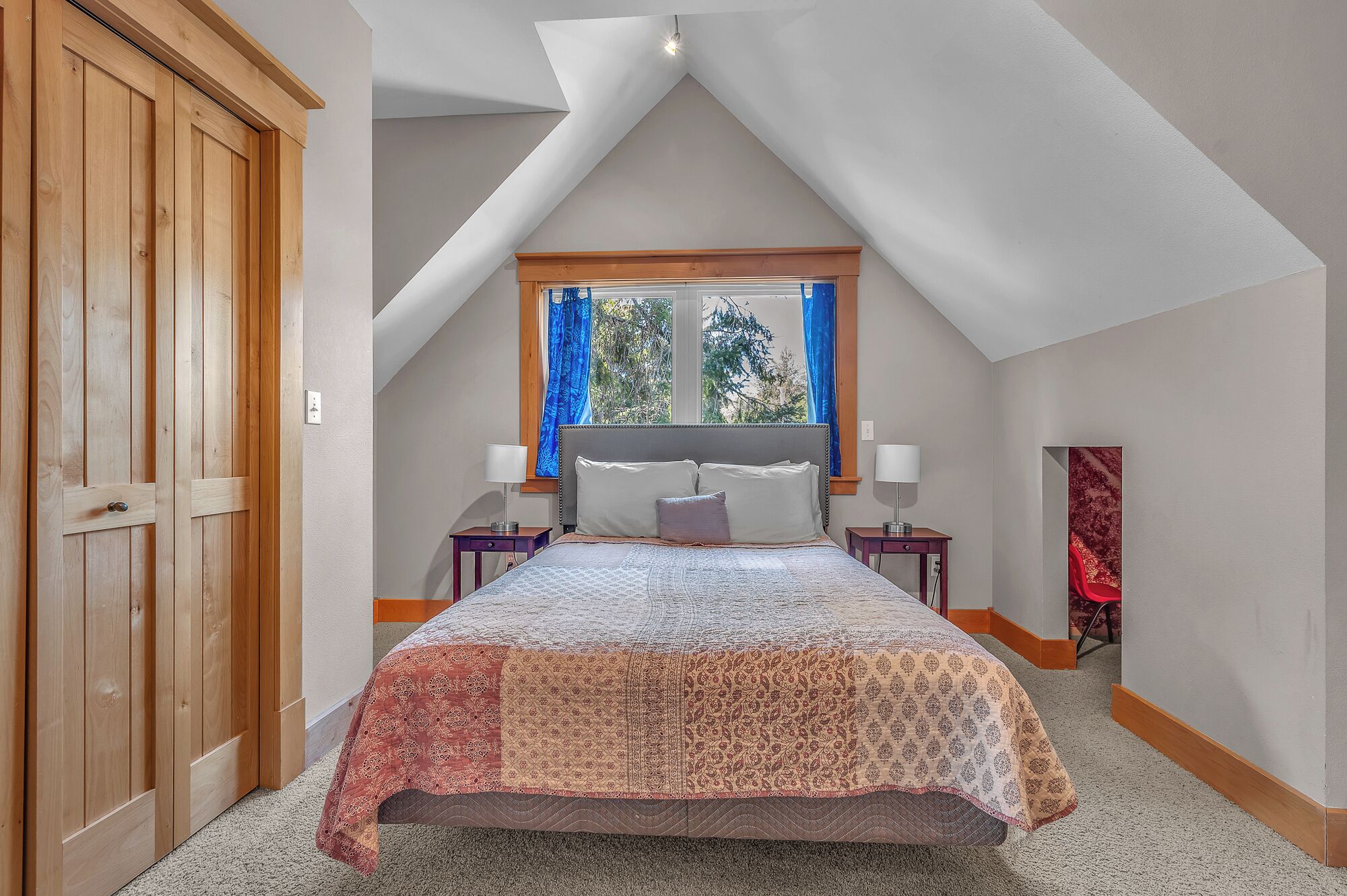 A bedroom with a sloped ceiling, a large bed with patterned bedding, two bedside tables with lamps, a window with curtains, and a door.