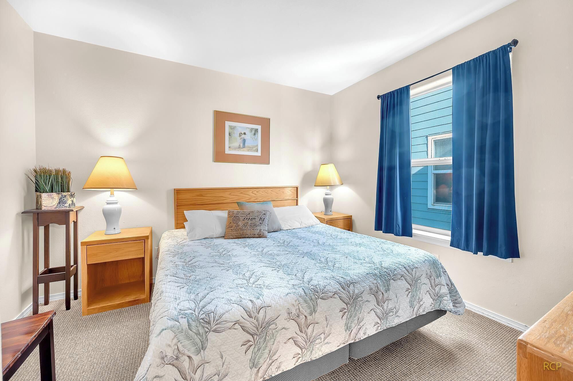 A bedroom with a bed, two bedside tables, a nightstand, a window with blue curtains, and a framed picture on the wall.