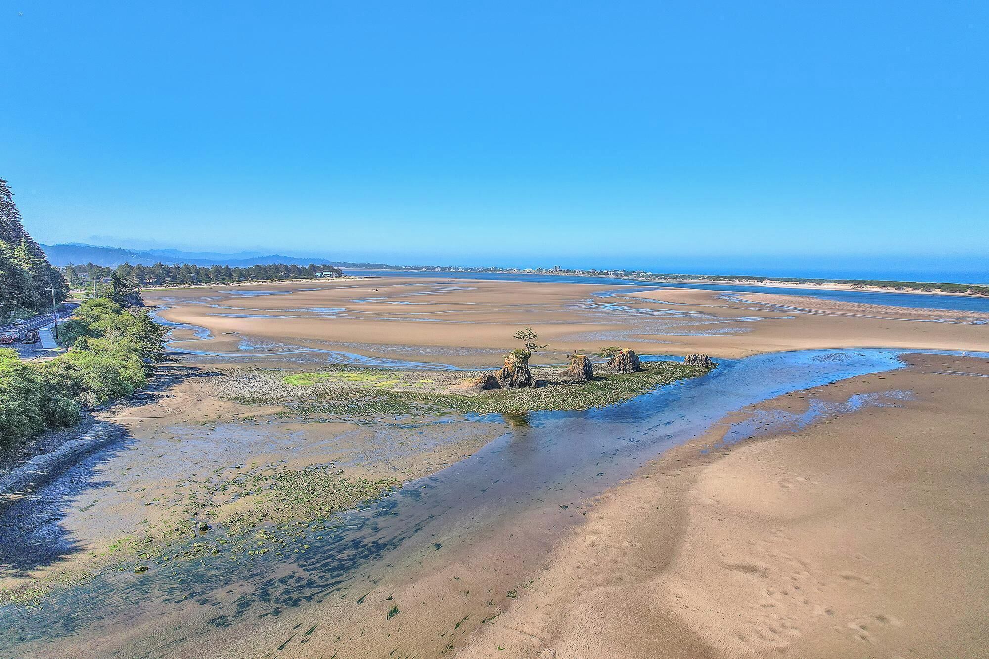 A coastal landscape with a wide sandy beach, a small rocky outcrop with vegetation, and a clear blue sky.
