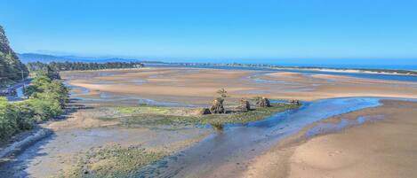 A coastal landscape with a wide sandy beach, a small rocky outcrop with vegetation, and a clear blue sky.