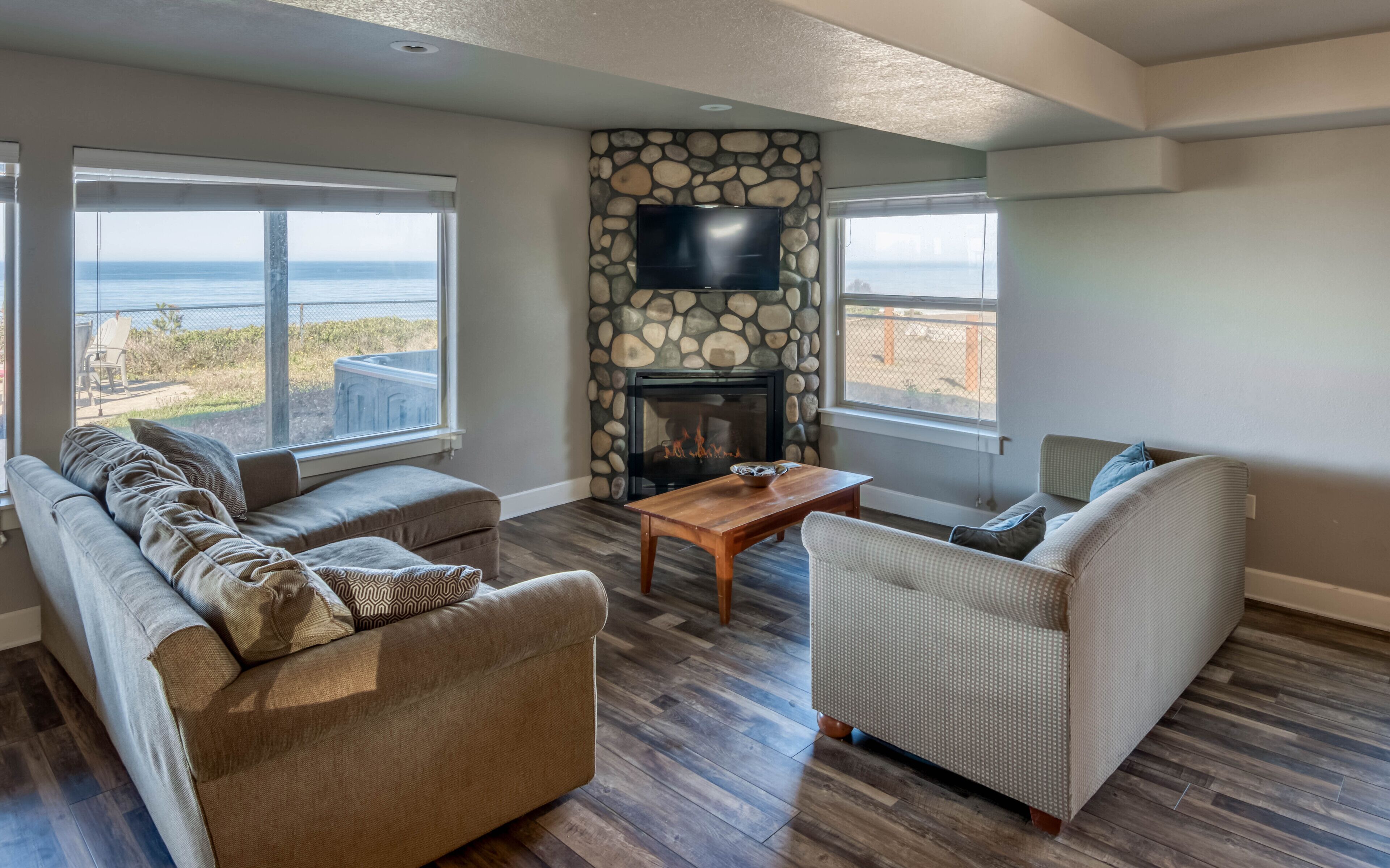 A living room with a stone fireplace, a flat-screen TV, and a view of the ocean.