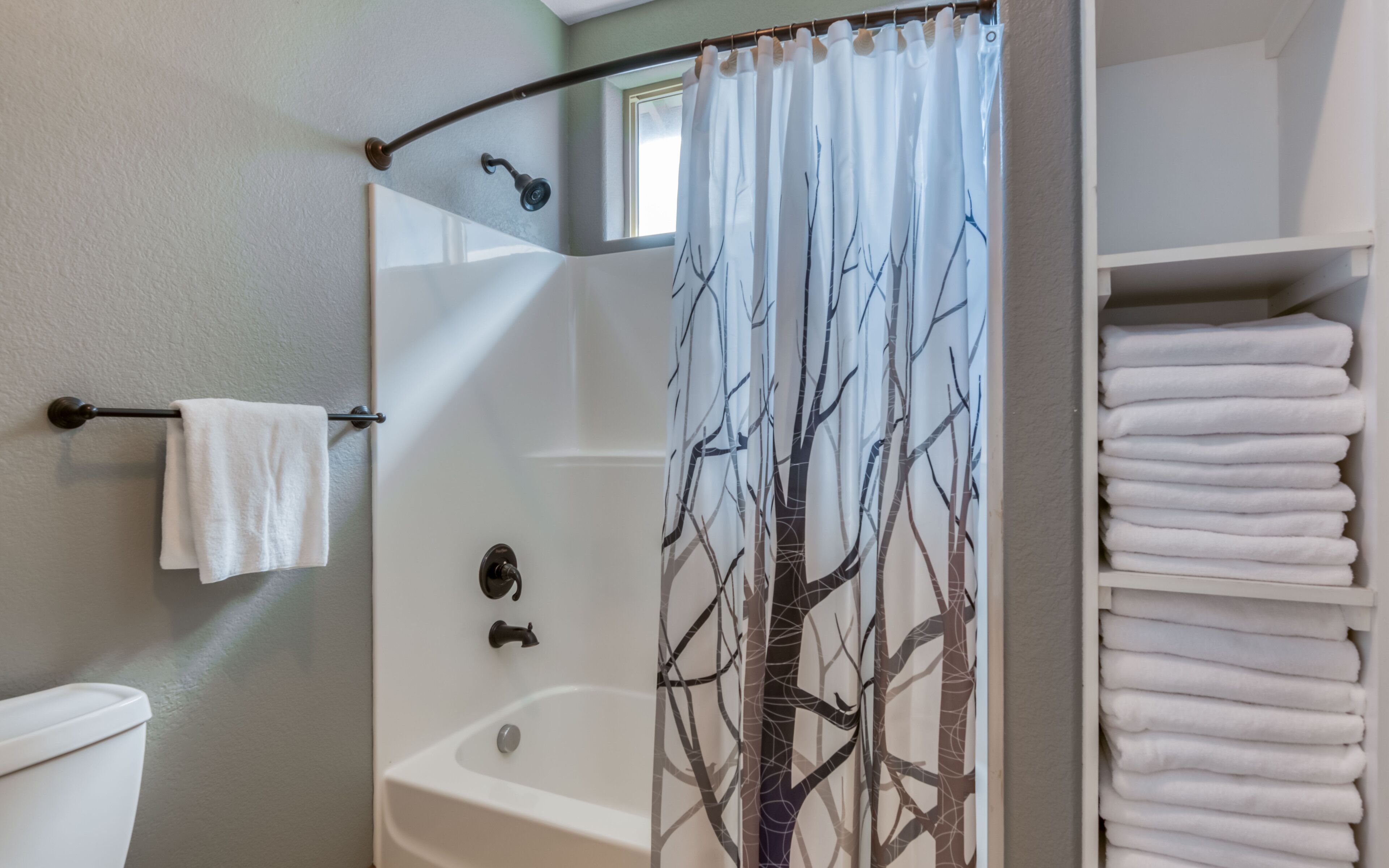 A bathroom with a shower curtain featuring a tree design, a bathtub, a toilet, and a stack of white towels.