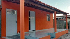 A single-story building with a red exterior and a tiled roof. It features two doors, one with a metal gate, and a small window. - Casa na praia do preá (Cruz)