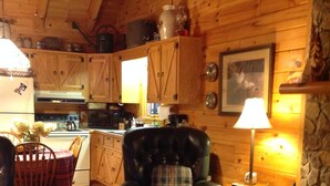 A cozy cabin interior with a dining area, a leather armchair, a rocking chair, and a kitchen with wooden cabinets.