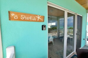 A teal wall with a wooden sign reading "8 Starfish" and a sliding glass door leading to a balcony with a view of the ocean. - The Starfish Oceanfront Studio Cottage in East End (East End)