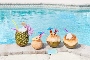 A poolside with four drinks in coconuts and a pineapple, and a small sign on the ground.