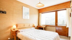 Une chambre à coucher avec une tête de lit en bois, un lit recouvert d’une couette blanche, une table de chevet avec une lampe, une fenêtre avec des rideaux et un luminaire fixé au mur.