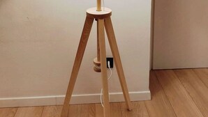 A floor lamp with a white shade on a tripod stand, positioned against a plain wall with a door to the right.