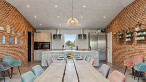 Una cucina moderna con isola centrale, un'area pranzo con un tavolo lungo e sedie colorate, e un muro in mattoni decorato con diversi cartelli.
