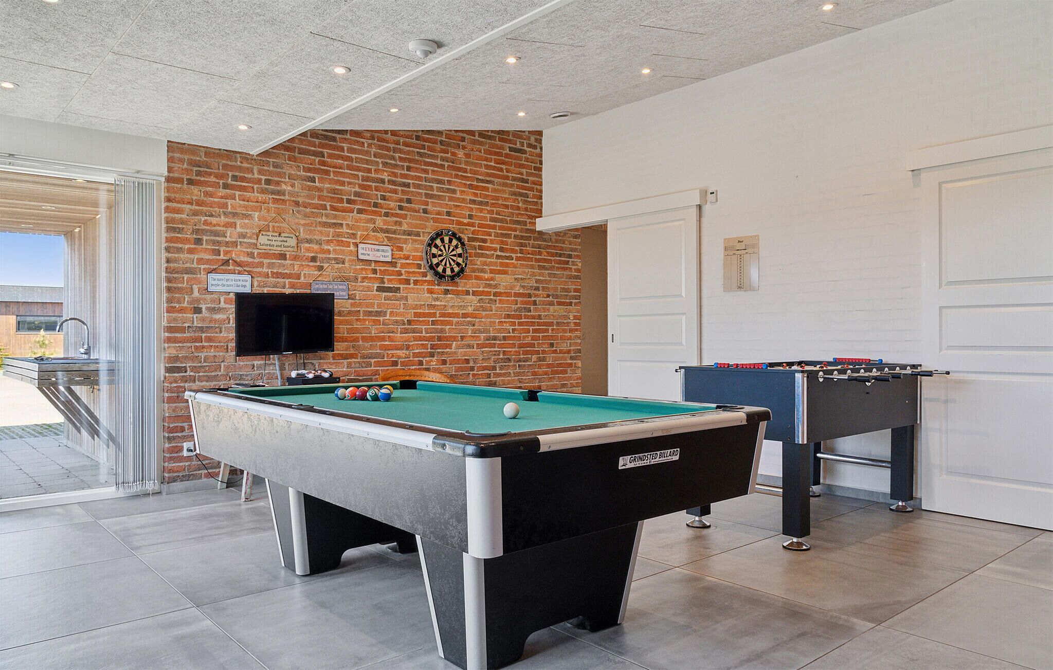Ein Billardtisch, eine Dartscheibe und ein Fußballtisch in einem Raum mit Backsteinwand und Tür.