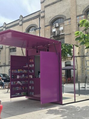 A purple kiosk with a book display, located on a sidewalk with a building in the background. - Alcantara 65 Riverside (Lisbon)