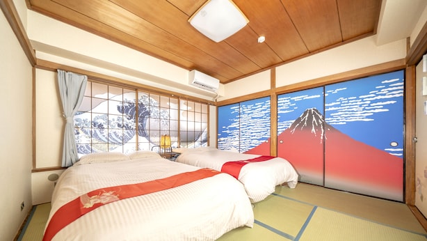Une pièce de style japonais traditionnel, avec un sol recouvert de tatamis, un lit avec une couvre-lit rouge et blanc, une fenêtre donnant sur une montagne et des portes coulissantes ornées d’une fresque représentant une montagne.