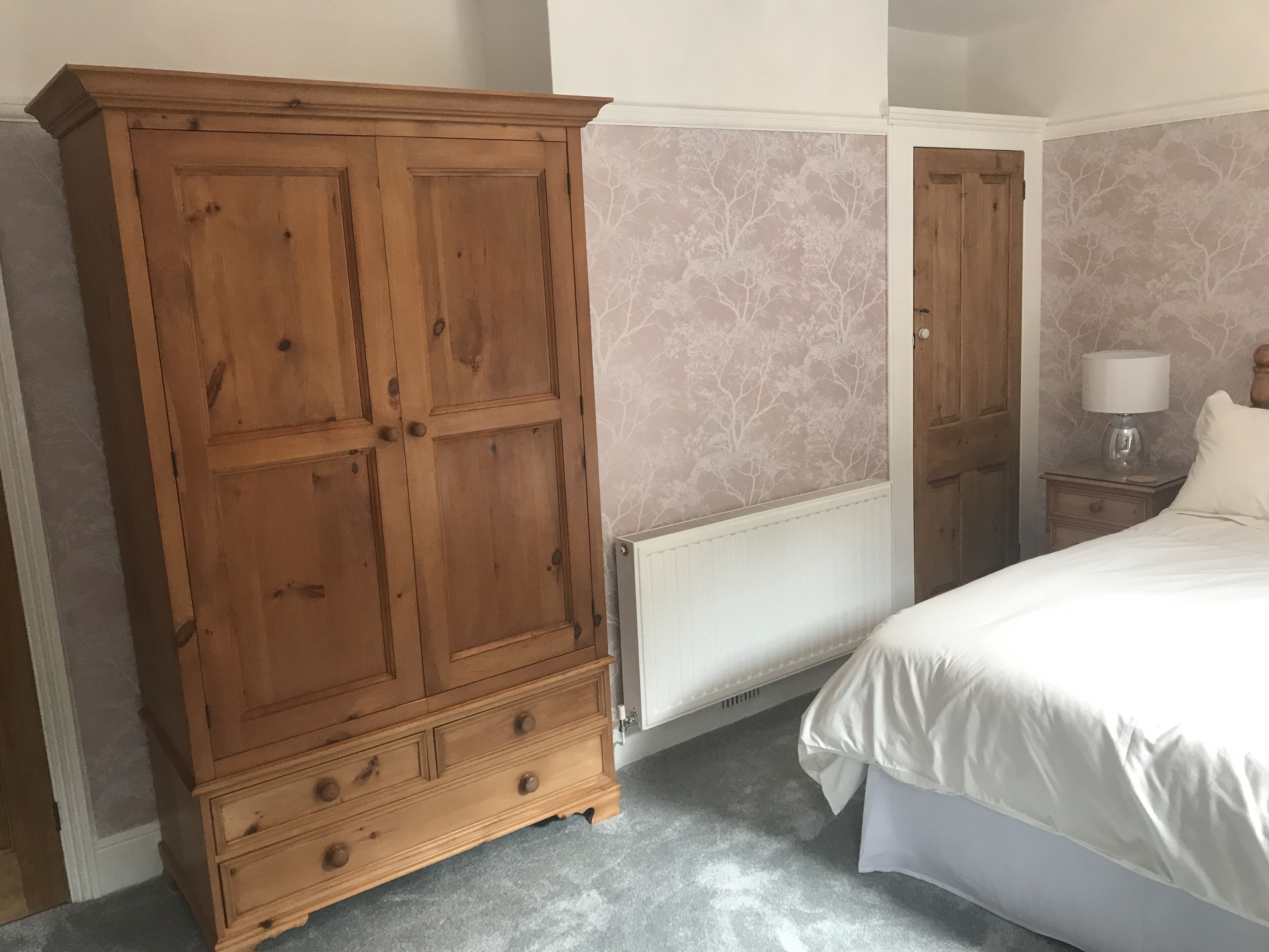 A bedroom with a large wooden wardrobe, a bed with white bedding, a nightstand with a lamp, and a radiator.