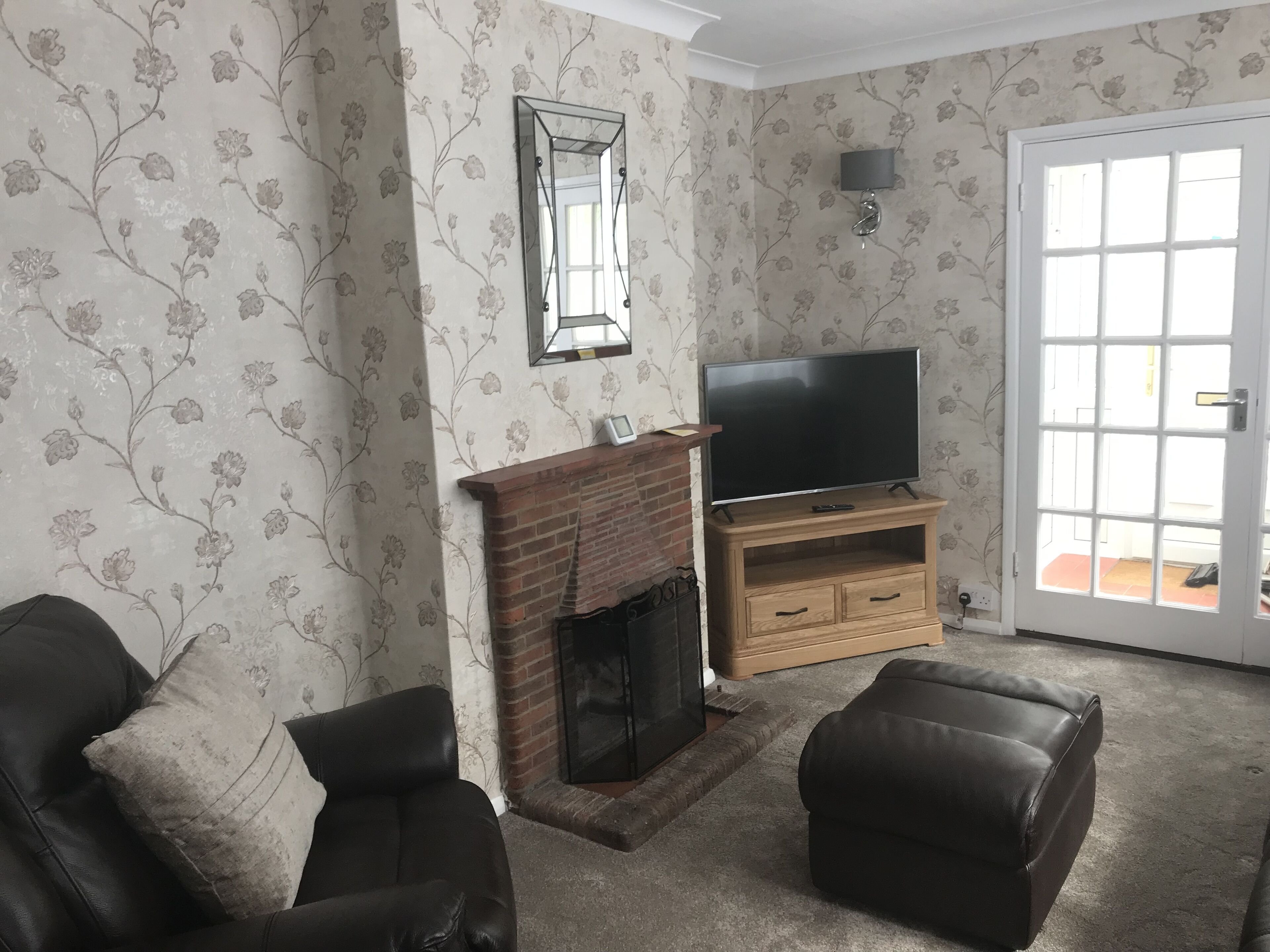 A living room with a fireplace, a television on a wooden stand, two leather armchairs, and a door with a view.