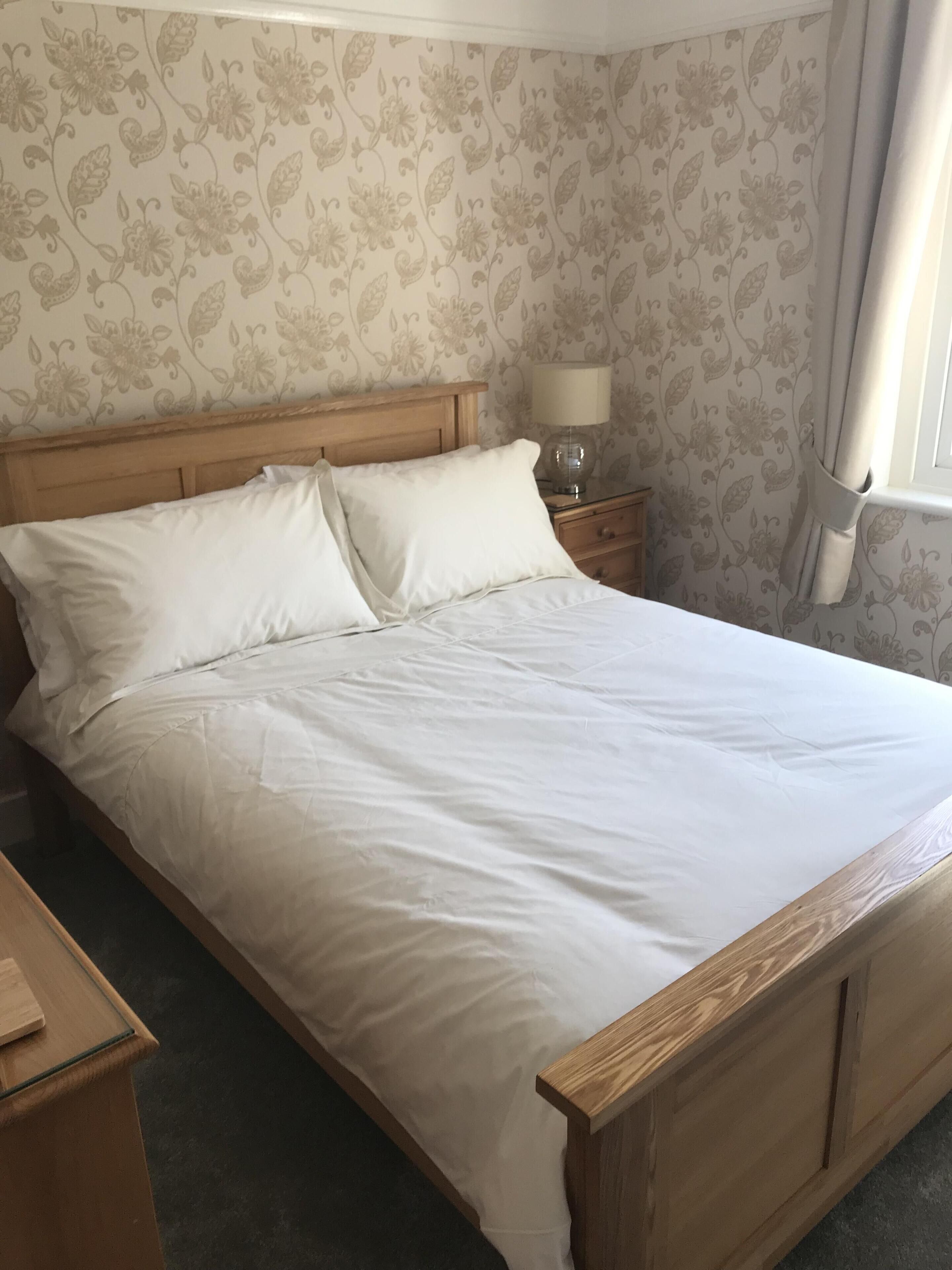 A bedroom with a wooden bed, white bedding, a nightstand with a lamp, and floral wallpaper.