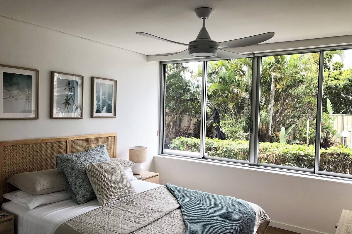 A bedroom with a bed, a nightstand, a lamp, and a ceiling fan. There is a window with a view of greenery outside.