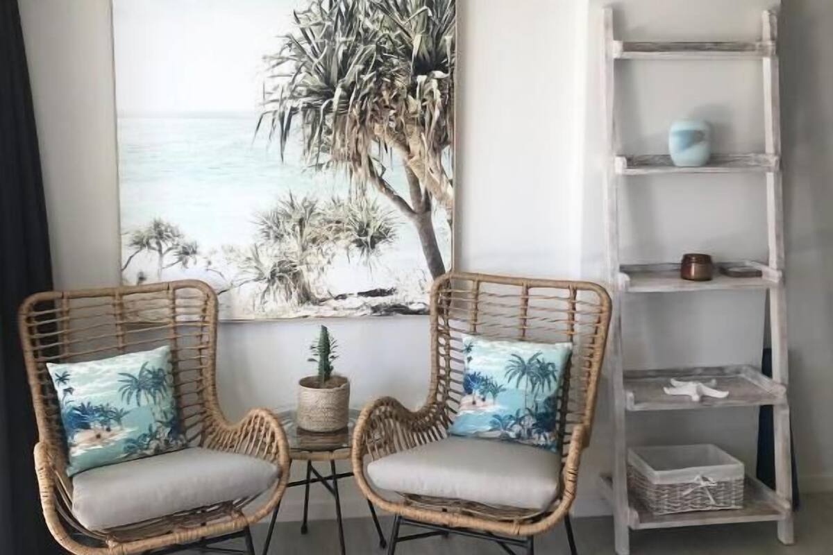 A room with two wicker chairs with cushions, a small table with a plant, and a white shelf with decorative items.