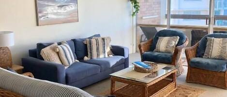 A living room with a sofa, armchairs, and a coffee table. There is a painting on the wall and a window providing natural light.
