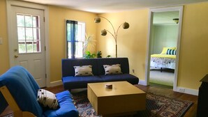 Un saló amb un sofà, una cadira de fusta, una taula de centre, una catifa amb dibuixos i una porta que dona a una altra habitació.