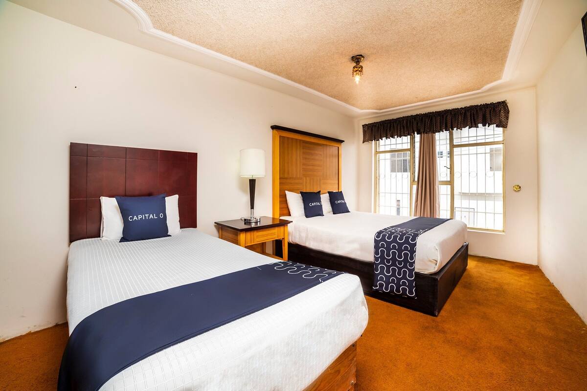 A hotel room with two beds, a wooden headboard, a nightstand with a lamp, and a window with curtains.