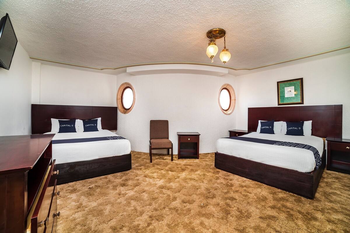 A hotel room with two beds, a chair, a small table, and a TV mounted on the wall.
