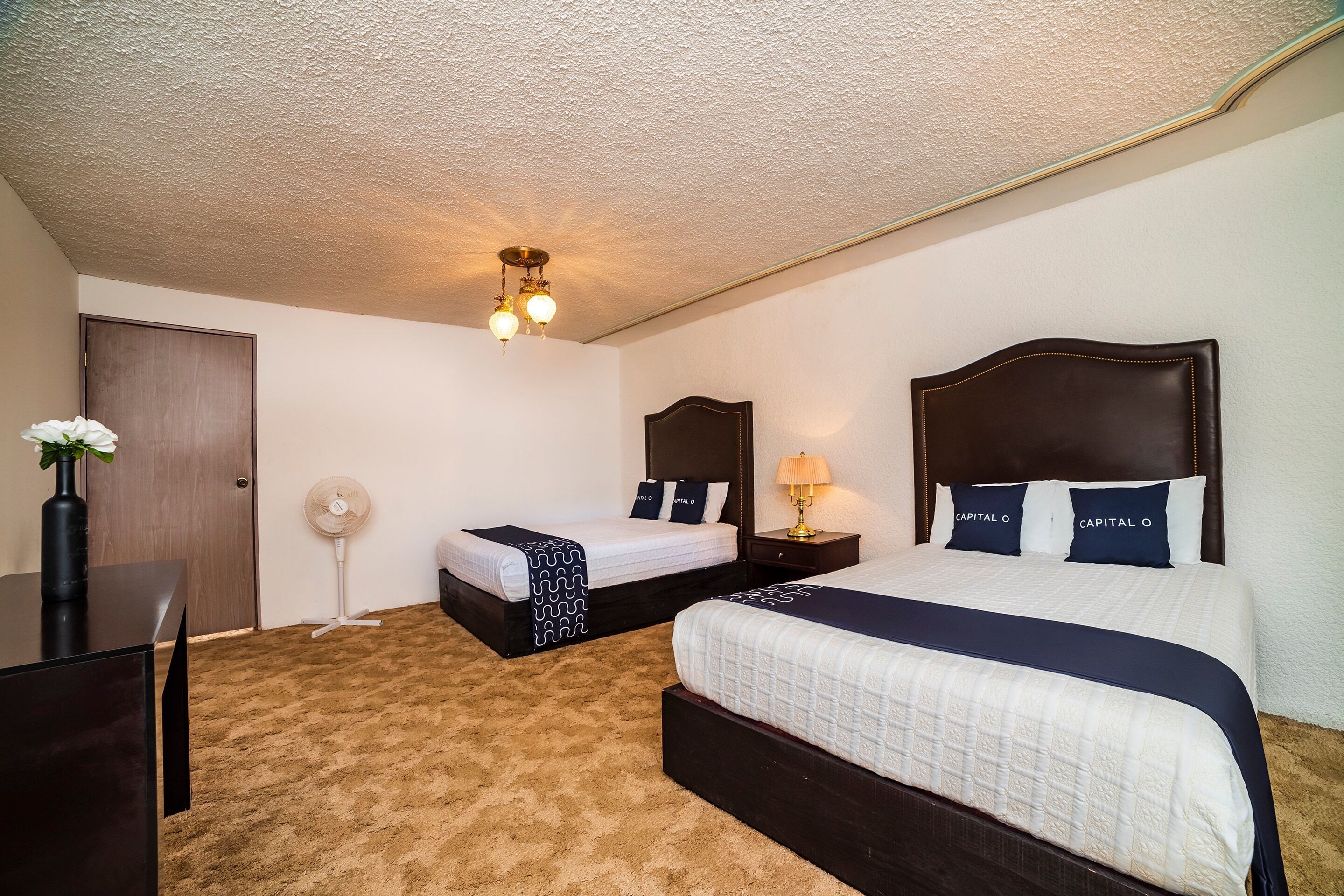 A hotel room with two beds, a desk, a chair, a dresser, a ceiling fan, and a wall-mounted lamp.