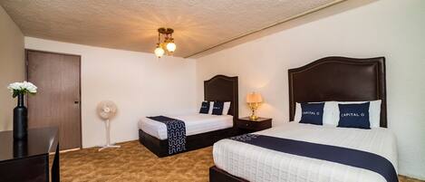 A hotel room with two beds, a desk, a chair, a dresser, a ceiling fan, and a wall-mounted lamp.