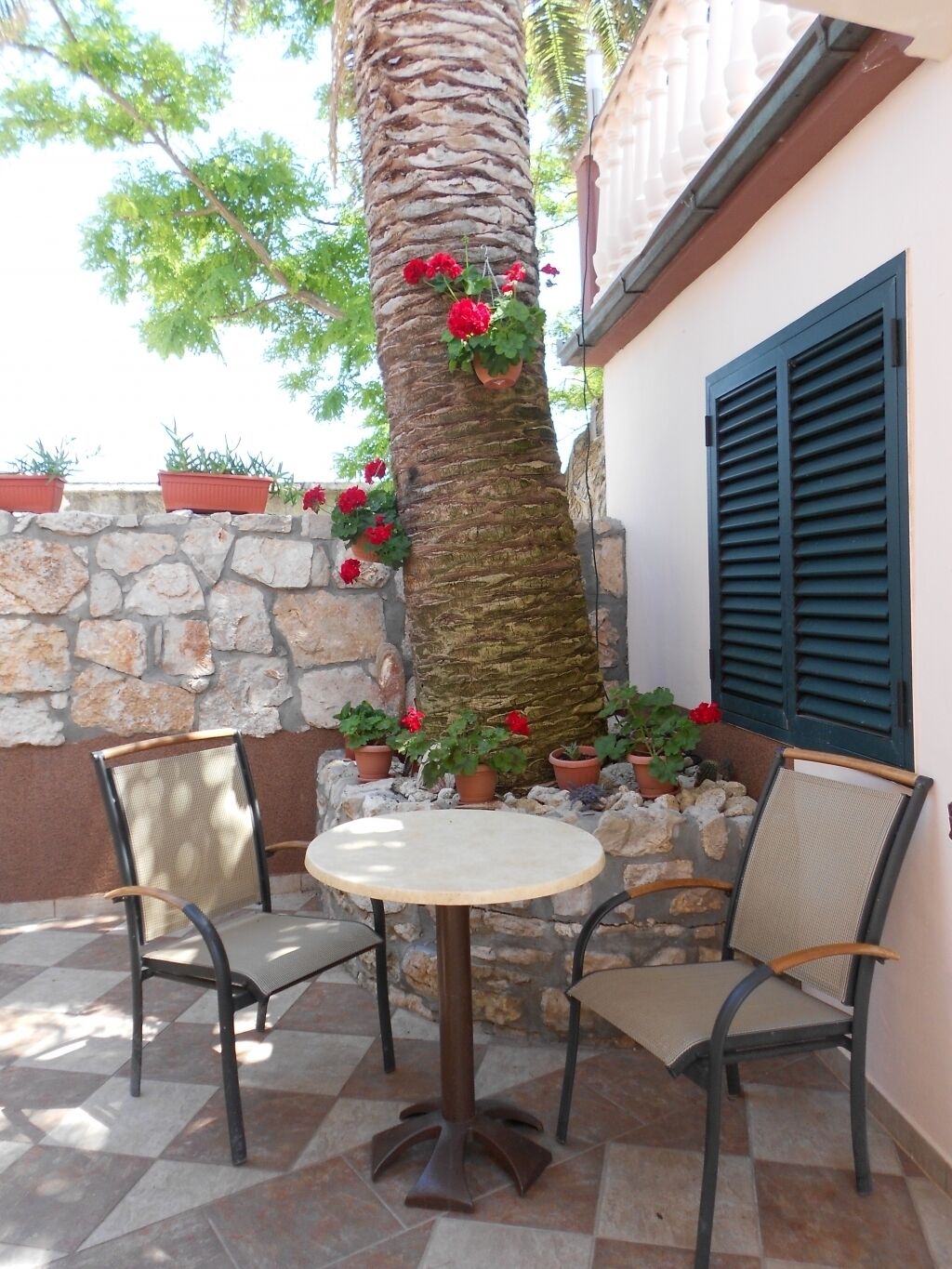 Un espace extérieur aménagé avec une table ronde et deux chaises, entouré de plantes en pot et d’un mur de pierres.