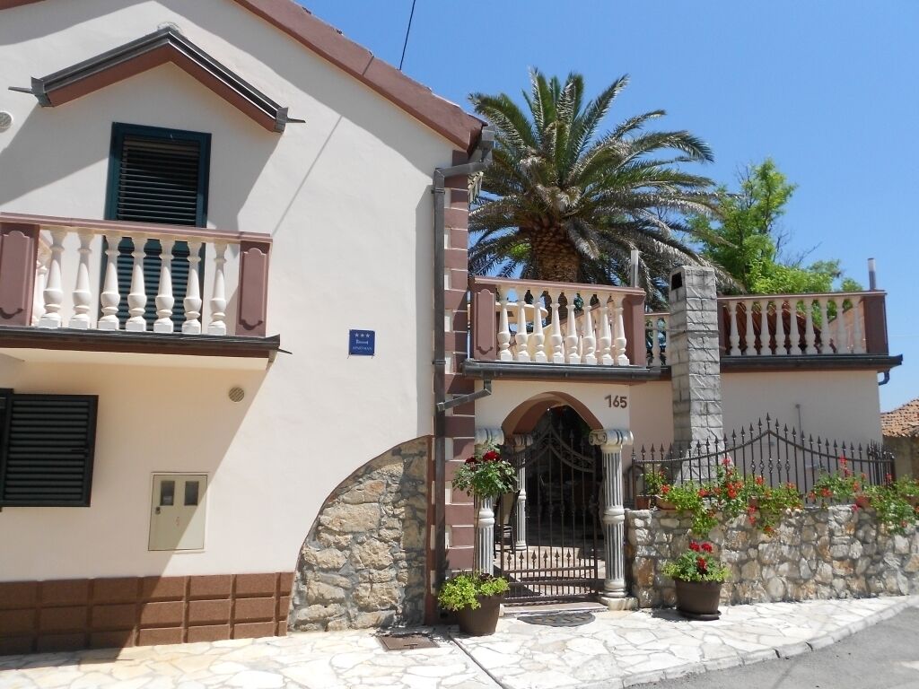 A building with a stone facade, a balcony with white balustrades, and a gate with a number '165' above it.