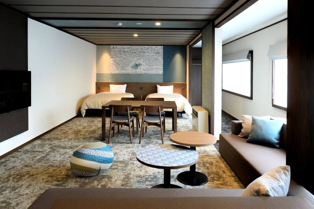 A modern hotel room with a sofa, two beds, a dining table, and a TV.